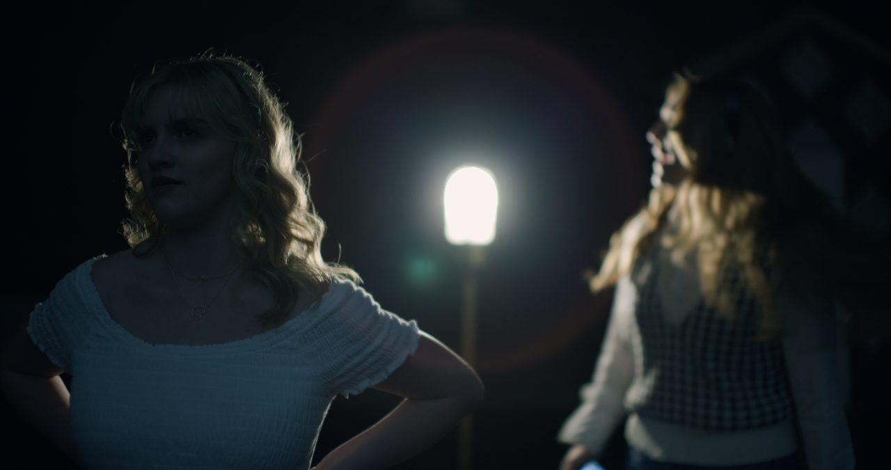Two women on a dark stage beside a glowing lamp