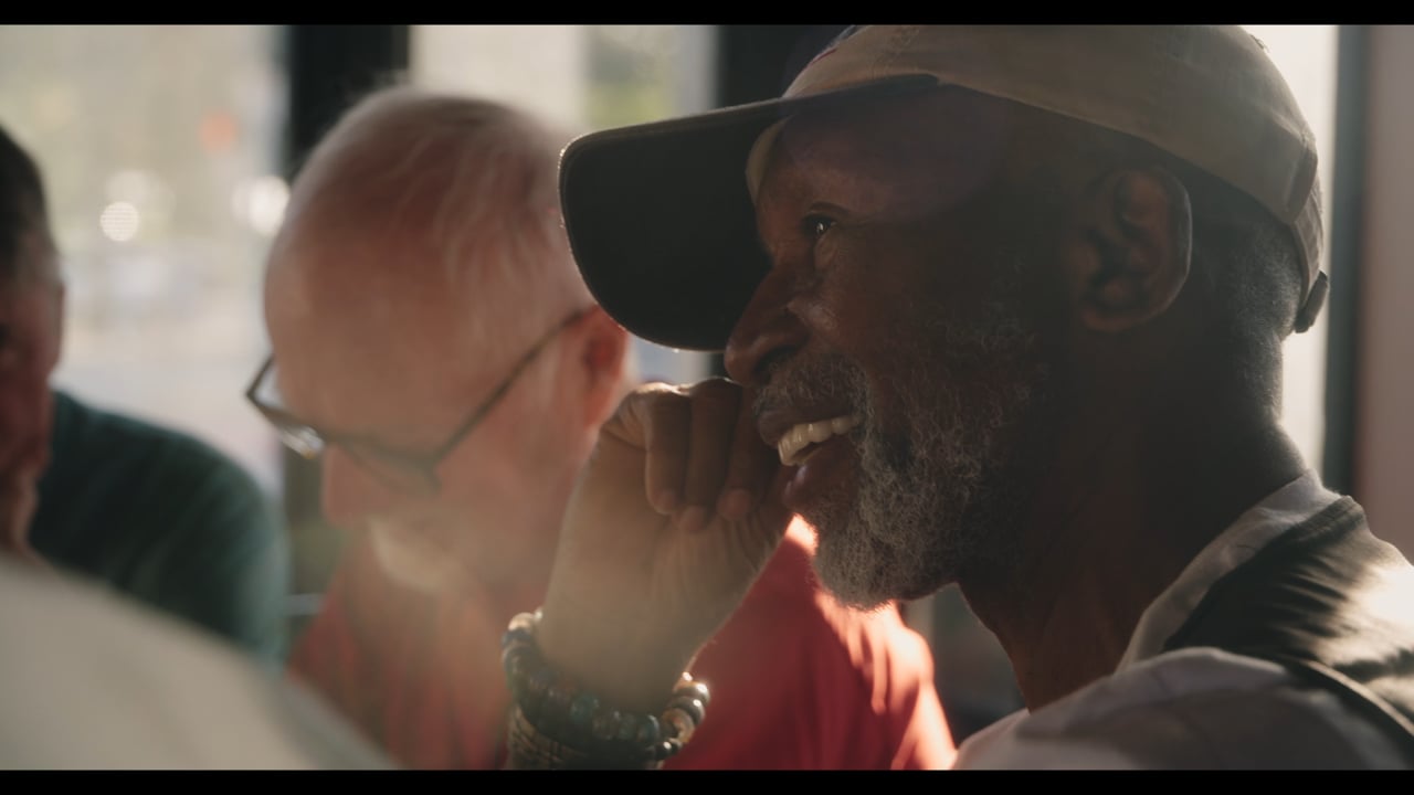 An older man in a cap smiles in warm window light beside friends